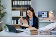 © Wasana - A woman is sitting at a desk with a laptop and a stack of papers