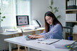 © Wasana - A woman is sitting at a desk with a laptop and a stack of papers