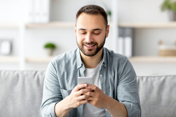 Poster - Positive young man sitting on couch with smartphone and texting to his friend, communicating online indoors. Millennial Caucasian guy writing sms, having video call or surfing web at home
