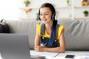 Poster - Modern Remote Learning Concept. Happy teen girl in headset studying online, sitting on sofa at desk, using laptop looking at screen, watching webinar, participating in lesson listening to teacher