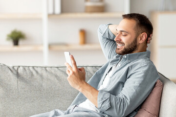 Poster - Handsome millennial guy relaxing on sofa with cellphone, smiling and texting his friend at home. Young Caucasian man writing message, having video chat or broswing internet at home