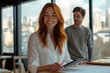 © Anjali - A smiling redhead woman reviews documents on a tablet, while a man stands in the background, in a modern office setting.