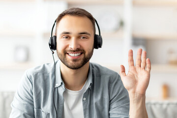 Poster - Headshot of smiling young guy wearing headphones, waving hand at webcam, greeting friend or colleague online, having video call or business conference, computer screen view