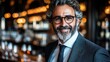 © JO BLA CO - A well-dressed man with curly hair and glasses stands confidently in a stylish bar, warmly smiling. The soft lighting creates an inviting atmosphere as patrons enjoy their drinks