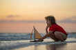© Volodymyr - kid traveling on sea. Kid playing with toy sailing boat, toy ship. Travel and adventure concept. Child dream of cruising. Kid on summer sunset beach. Kid playing on the beach at the sunset.