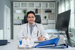 © Kawee - Portrait of Asian mature male doctor sitting on table in hospital office.