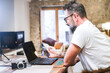 © simona - Middle-aged man sitting at his home office desk using laptop and smartphone, working remotely on his freelance photography business, with a camera on the table and fireplace in the background
