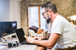 © simona - Middle-aged man sitting at home office desk working on laptop with camera on table, living room background with sofa and fireplace, concept of remote work, digital profession, modern workspace
