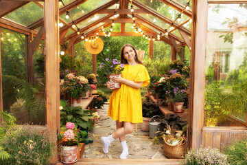 Poster - Funny gardener girl with plants at backyard home. Pretty teen girl in yellow dress holding with flower in pot in  orangery. Young girl holding houseplant on backyard in greenhouse with flowers in pots