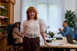 © DusanJelicic - Redhead businesswoman is posing in a modern office with hands in pockets while her team is working in the background, demonstrating leadership and teamwork in a collaborative workspace