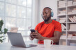 © Tj - Young black man wearing headphones and using smartphone while working on a laptop at home, with a cup of coffee on the table and bookshelf in the background