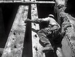 © Kulvarin - A worker climbs wooden beams during construction, showcasing labor and determination in a vintage black-and-white setting.
