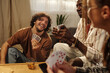 © TrueFrame Collective - Young cheerful man with fan of playing cards sitting in front of intercultural couple during game