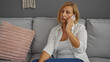 © Krakenimages.com - Middle-aged woman with short blonde hair talking on the phone in a home living room setting, seated on a gray couch with a pink pillow, wearing a white shirt and jeans, appearing thoughtful and calm.