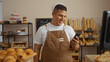 © Krakenimages.com - Young hispanic man in a bakery shop looking at his phone while smiling, wearing a brown apron, surrounded by various types of bread and pastries on display in an inviting indoor setting.