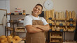 © Krakenimages.com - Smiling young hispanic man with crossed arms in a bakery, surrounded by fresh bread and pastries, standing confidently in an indoor latin shop setting.