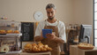 © Krakenimages.com - Young man with a beard in a bakery shop using a tablet, surrounded by croissants and pastries, with a clock and a chalkboard menu in the background.