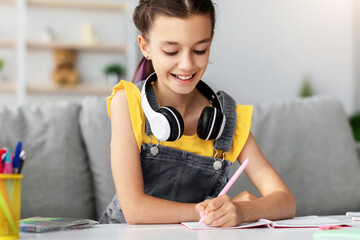 Poster - Lifestyle Concept. Smiling school girl with colorful dyed hair sitting at desk, writing essay in copybook. Pupil with headset studying and doing homework or taking notes in her diary, making checklist