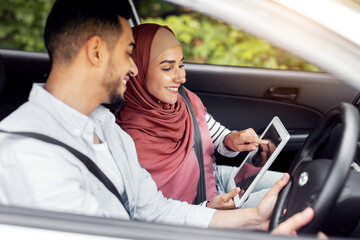 Cheerful millennial muslim female in hijab shows way on tablet with empty screen to husband at steering wheel in car, outdoor. Digital map, online application for travel, device and road to vacation