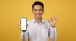 © mamo studios - Smiling handsome young Asian man wearing a white checkered shirt showing a mobile phone with a blank screen and okay sign isolated over yellow background. People Lifestyle Concept