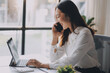 © ARMMY PICCA - Young asian woman talking on mobile phone while taking notes and working with laptop computer at home. Business, home office and technology concept.