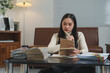 © Phimwilai - Focused young woman in a modern office setting, analyzing documents and taking notes. Papers and files are spread on the table, indicating a busy work environment
