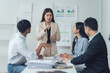 © Kritdanai - Business Strategy Session: A diverse team of business professionals engaged in a lively discussion around a table, with a female leader guiding the group.