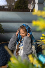 A beautiful Russian girl in a fur coat and headscarf sits on the porch of a wooden house on a sunny spring day.Family values,traditions and home comfort.The girl covers her hair with a beautiful scarf