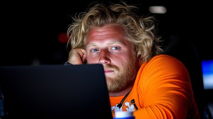 Poster - Man with a beard and long hair is sitting in front of a laptop. He is wearing an orange shirt and he is tired