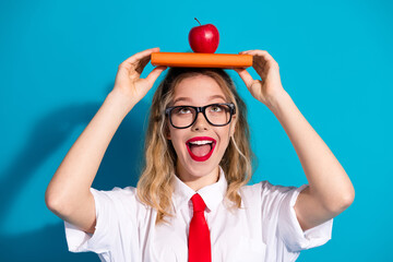 A vibrant young woman balances a book with an apple on her head, displaying a fun academic theme against a vibrant blue backdrop.