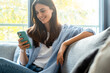 © simona - Young millennial woman sitting on a couch at home, casually dressed, smiling while using a smartphone to browse the internet, scroll social media, text friends, subscribe to online services
