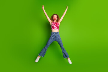 Wall Mural - Joyful young woman jumping with arms raised on vibrant green background showcasing casual style