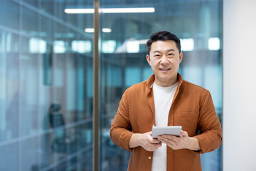 Canvas Print - Portrait of successful Asian man inside office. Businessman smiling and looking at camera while holding tablet computer. Programmer developer at workplace near window.