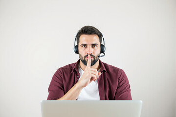 Aufkleber - Thoughtful man with headset gesturing silence while working on laptop against light background.