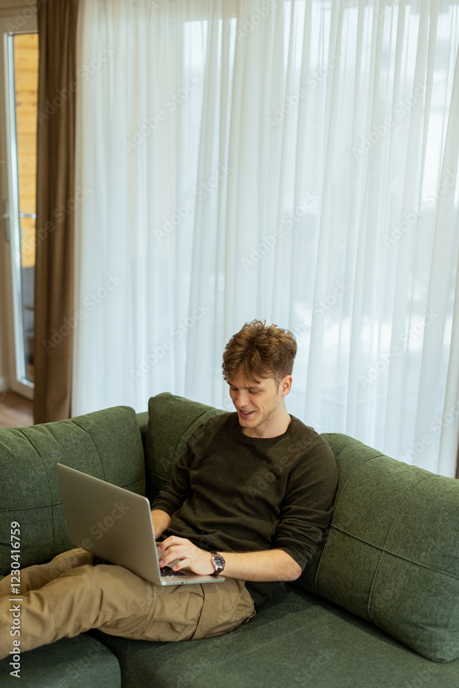 Man working diligently on his laptop while relaxing on a cozy green couch in a bright, airy living room