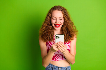 Wall Mural - Cheerful young woman with curly red hair using a smartphone against a green backdrop showcasing a stylish and vibrant lifestyle