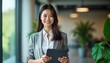 © Vadym - Young Asian businesswoman stands in modern office holding tablet. Pro woman looks happy, confident. Planning working on something. Modern tech office environment with plants. Possible scenario for