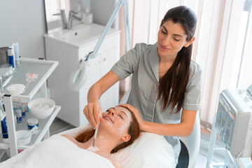 Wall Mural - Professional cosmetology: Smiling massage therapist applying face mask to woman with brush at salon. From above of relaxed female customer is getting pampered in spa by beautician