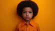 © imagineRbc - A young child stands confidently with an intense gaze, wearing an orange shirt. The bright yellow background enhances the child's unique hairstyle and serious demeanor, highlighting individuality