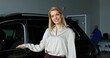 © m-art - A stylish woman smiles looking at the camera standing next to a car in a car dealership
