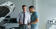 © m-art - A car dealership manager talks to a customer standing next to a car with the hood open in the showroom