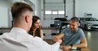 © m-art - A man signs a car purchase agreement at a car dealership. The manager is sitting at his desk in front of an open laptop