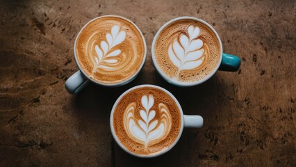Poster - Three elegant latte art designs in white on warm brown coffee cups arranged in a triangular formation over a textured wooden table surface.
