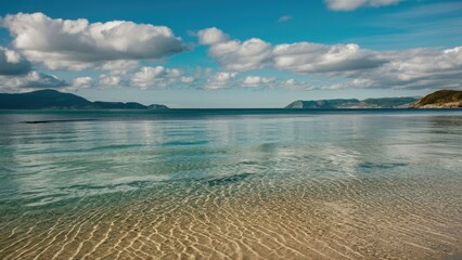 Aufkleber - Serene beach landscape with clear blue water, gentle ripples reflecting clouds, and distant mountains under a bright sky in tranquil coastal setting