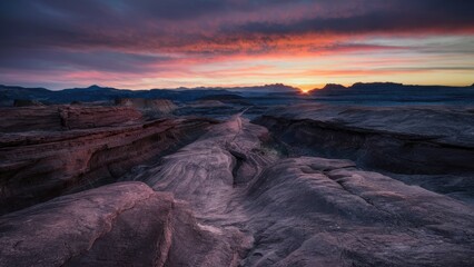 Poster - Majestic sunset over rocky landscape with purple and orange hues illuminating rugged terrain and distant mountains under a dramatic sky.