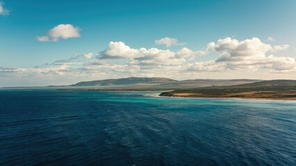 Poster - Vast deep blue ocean under a clear sky with soft white clouds, showcasing a panoramic view of coastal landscape and rolling hills in background