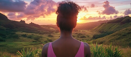 Wall Mural - backview of a woman with curly hair in a pink top observing a vibrant sunset over a lush green valle
