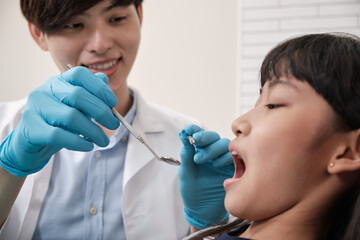 Wall Mural - A young Asian male pediatric dentist checks and examines a little Thai girl's teeth in dental clinic, well-being mouth oral hygiene, and professional orthodontic healthcare work in a kid hospital.