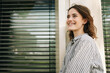 © contrastwerkstatt - Young woman smiling in front of window blinds