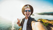 © Davide Angelini - Young man wearing hat and sunglasses taking selfie picture with cellphone on the top of the hill - Traveler male person with backpack smiling at camera at the sea